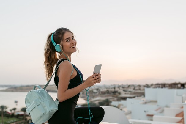 Genieten van heerlijke muziek via een koptelefoon van een positieve, vrolijke sportvrouw die bij zonsopgang aan het strand chillen. Modieus model, plezier maken., Glimlachend.