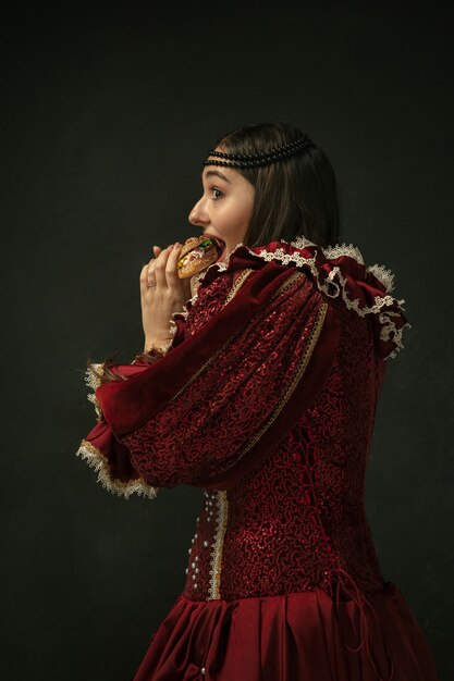 Gepassioneerd. Portret van middeleeuwse jonge vrouw in rode vintage kleding hamburger eten op donkere achtergrond. Vrouwelijk model als hertogin, koninklijk persoon. Concept vergelijking van tijdperken, modern, mode, schoonheid.