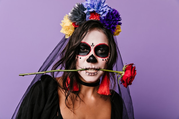 Gepassioneerde Mexicaanse vrouw met geverfd gezicht met rode roos in haar tanden. Close-upfoto van krullende brunette met kleurrijke bloemen in haar haar.