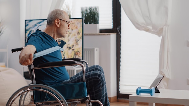 Gepensioneerde man die in een rolstoel zit en weerstandsband gebruikt om te oefenen terwijl hij de trainingsvideo op tablet volgt. Oudere volwassene met lichamelijke handicap die elastische riem trekt om uit te rekken