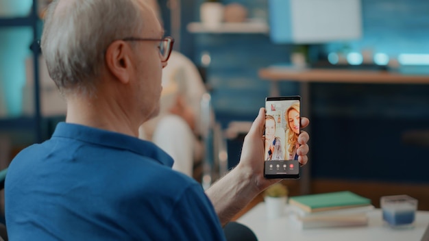 Gepensioneerde man praat met familie op online videoconferentie, met mobiele telefoon voor communicatie op afstand. Oudere persoon die internetteleconferentie op smartphone gebruikt om met familieleden te chatten.