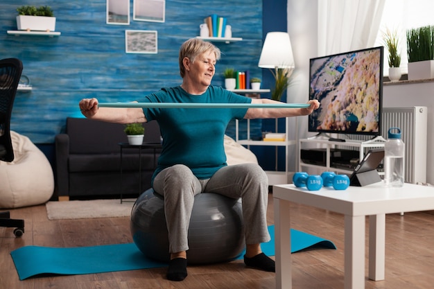 Gepensioneerde senior man zit op fitness zwitserse bal in woonkamer wellness fitness workout streching armspieren te doen met behulp van aerobics elastische band