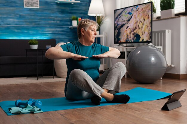 Gepensioneerde senior vrouw eet fitness tutorial op laptop zittend op yoga mat strekkende arm
