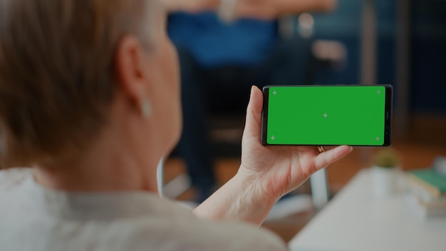 Gepensioneerde volwassene die smartphone met horizontaal groen scherm thuis vasthoudt. Senior vrouw die geïsoleerde mock-up sjabloon analyseert en ruimteachtergrond kopieert met lege Chromakey op mobiele telefoon.