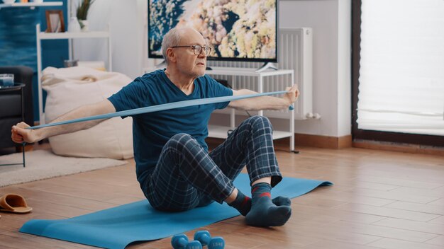 Gepensioneerde volwassene die weerstandsband trekt en thuis op yogamat zit. Volwassen persoon die rekoefeningen doet met flexibele elastische riem om de armspieren te trainen. Gepensioneerde opleiding