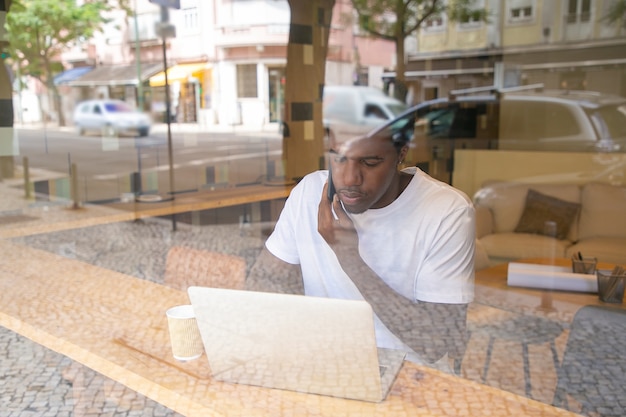 Gerichte Afro-Amerikaanse ondernemer die op laptop werkt en op mobiel in co-working space spreekt