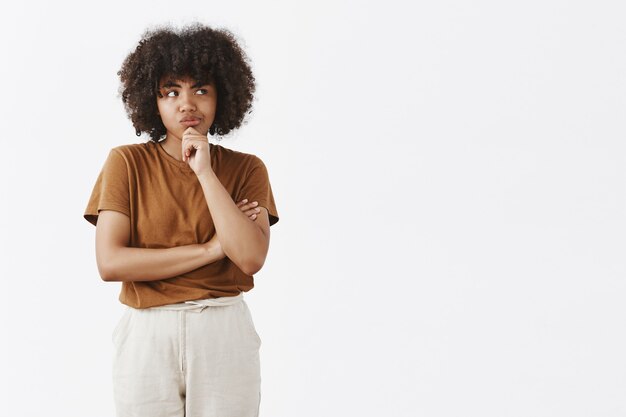 Gerichte doordachte schattig meisje met afro kapsel in bruin t-shirt en broek grijnzend op zoek naar rechts terwijl denken met twijfels