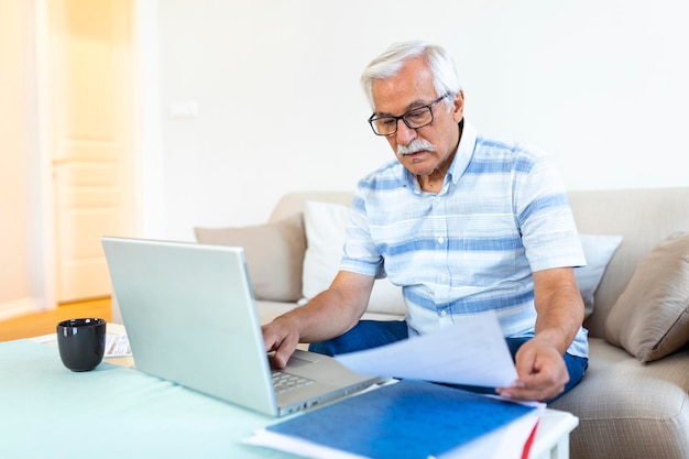 Gerichte grijsharige oudere man zit op de bank bankmeldingen te lezen en binnenlandse uitgaven te berekenen geconcentreerde moderne volwassen man overweegt financiële administratie om rekeningen op laptop online te betalen