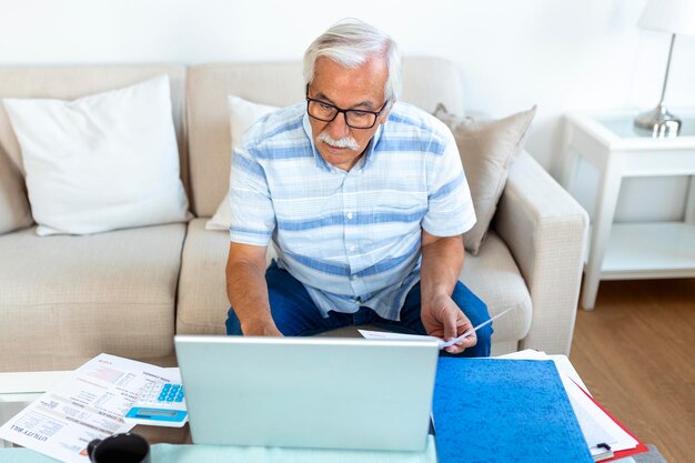 Gerichte grijsharige oudere man zit op de bank bankmeldingen te lezen en binnenlandse uitgaven te berekenen geconcentreerde moderne volwassen man overweegt financiële administratie om rekeningen op laptop online te betalen