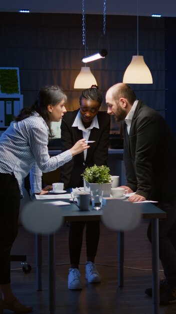 Gerichte leidervrouw komt 's avonds laat in de kantoorvergaderruimte leunend op de vergadertafel brainstormen over de bedrijfspresentatie. Diverse multi-etnische teamwerk oplossende managementstrategie