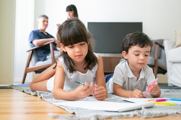 Gerichte schattige kleine broer en zus liggend op de vloer en tekening in de woonkamer terwijl ouders samen zitten