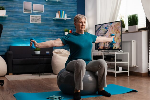 Gerichte senior vrouw strekkende arm werken aan lichaamsspier met behulp van fitness halters zittend op de Zwitserse bal in de woonkamer. Kaukasisch mannetje dat gespierde gezondheidszorg uitoefent tijdens wellnesstraining