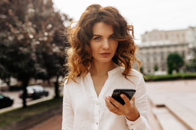 Gerichte stijlvolle vrouw met krullend haar SMS-bericht in herfstdag Mooie vrouw wandelen door groene struiken met een glimlach