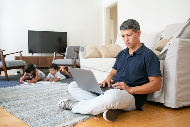 Gerichte vader thuis werken, zittend op de vloer en met behulp van de computer terwijl zijn kleine kinderen tekenen