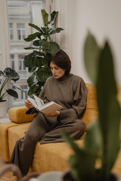 Gerichte volwassen blanke vrouw die een boek over tuinieren leest terwijl ze op de bank zit in een lichte kamer. Concept van tijd doorbrengen met hobby