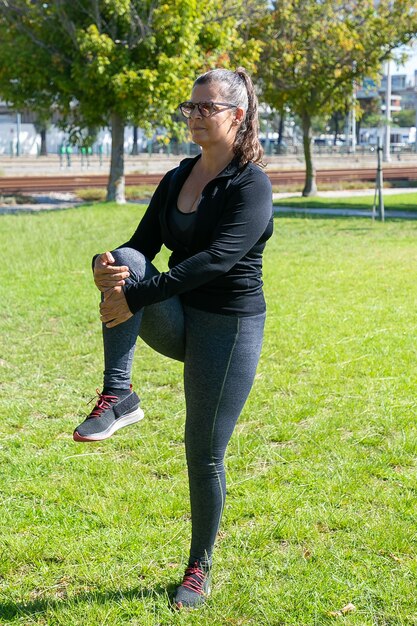 Gerichte volwassen vrouw in fitness kleding ochtend oefening op park gras, staande en benen strekken. Welzijn of actief levensstijlconcept