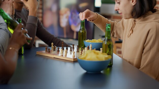 Gerichte vrouw die strategische bordspellen speelt met vrienden, plezier heeft met schaakwedstrijd en competitie thuis. Gelukkige mensen die samen genieten van gameplay, bier serveren. Uit de hand geschoten. Detailopname.