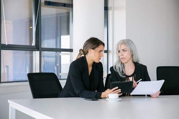 Gerichte vrouwelijke collega's die rapporten bespreken en analyseren. Twee professionals zitten samen, houden documenten vast, gebruiken tablet en praten. Teamwerk concept