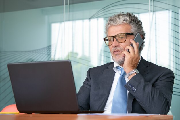 Gerichte zakenman in pak en bril bespreken deal op mobiele telefoon, werken op laptop in kantoor, display kijken