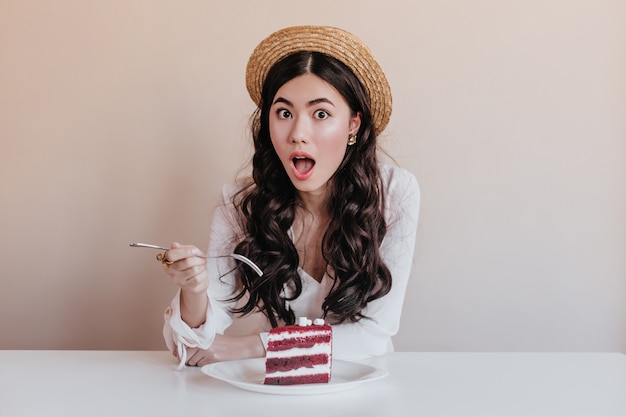 Geschokt Aziatische vrouw die in hoed dessert eet. Verbaasde Chinese vrouw die van cake geniet.