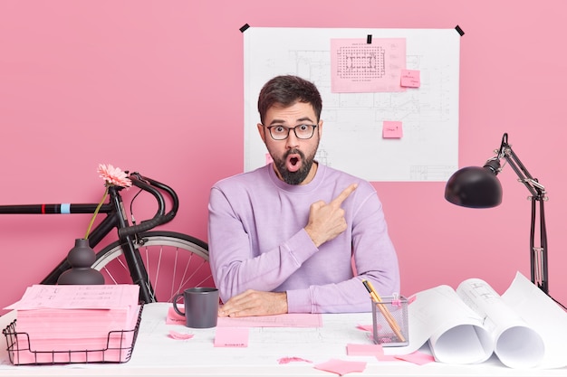 Geschokt bebaarde man kantoormedewerker wijst op roze muur demonstreert schetsen poses op desktop schrijft informatie op stickers heeft afstand baan zit op coworking space