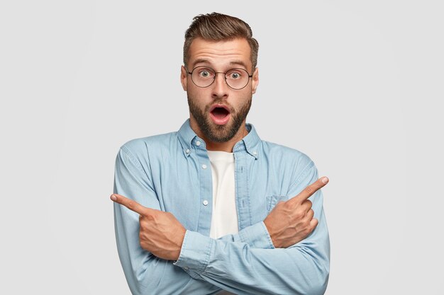 Geschokt blanke man wijst aan verschillende kanten met wijsvingers, kan niet kiezen tussen twee items, heeft een verbaasde uitdrukking, draagt een ronde bril en een blauw shirt, geïsoleerd over een witte muur