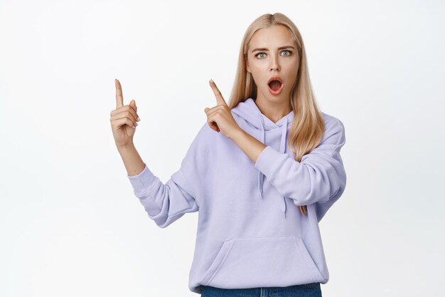 Geschokt blond meisje dat met de vingers naar de linkerbovenhoek wijst, laat de kaak vallen en staart onder de indruk naar de camera die in een hoodie staat tegen een witte achtergrond