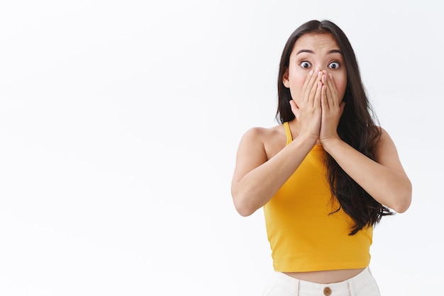 Geschokt en bezorgd jonge vrouw hijgend van angst of medelijden als iemand gekwetst is, bedek de geopende mond en staar camera nerveus angstig zie accdent gebeurt witte achtergrond Emoties concept