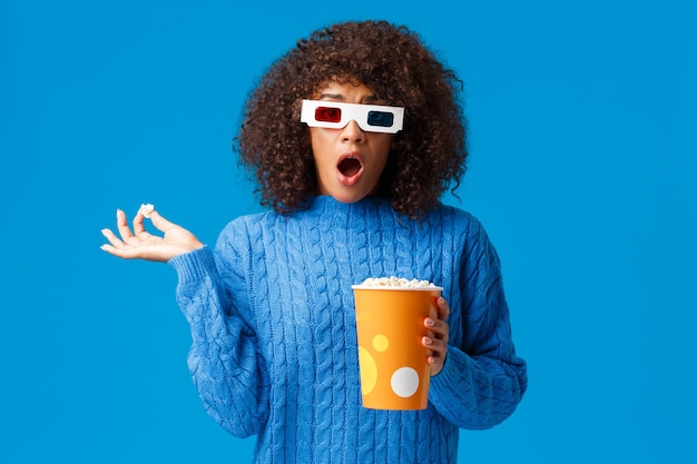 Geschokt en geschrokken jong schattig afrikaans-amerikaans hipstermeisje dat naar actiefilm kijkt, drama naar adem snakkend en kaak laat vallen terwijl ze popcorn eet en een schokkende scène in film ziet met 3d-bril, blauwe achtergrond.