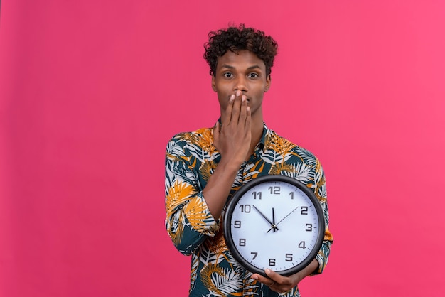 Geschokt en verward jonge donkerhuidige man met krullend haar in bladeren bedrukt overhemd met wandklok met tijd met handen voor mond op een roze achtergrond