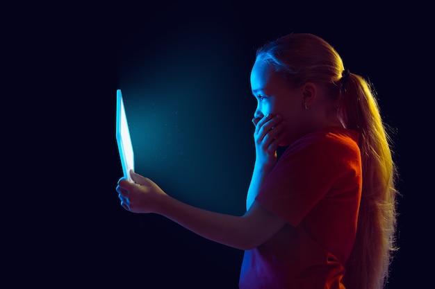 Geschokt. Het portret van het Kaukasische meisje dat op donkere studioachtergrond in neonlicht wordt geïsoleerd. Mooi vrouwelijk model dat tablet gebruikt. Concept van menselijke emoties, gezichtsuitdrukking, verkoop, advertentie, moderne technologie, gadgets.