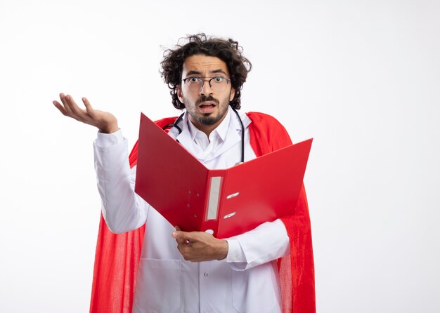 Geschokt jonge blanke man in optische bril dragen arts uniform met rode mantel en met een stethoscoop om de nek staat met opgeheven hand en houdt de map met kopie ruimte