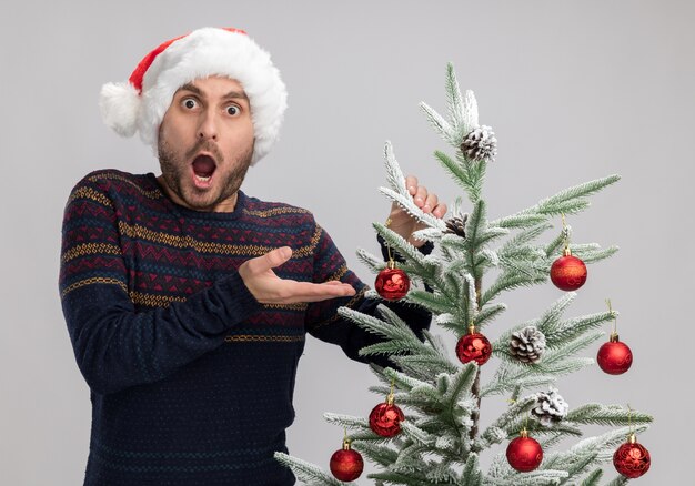 Geschokt jonge blanke man met kerstmuts staande in de buurt van de kerstboom aan te raken kijken naar camera geïsoleerd op een witte achtergrond