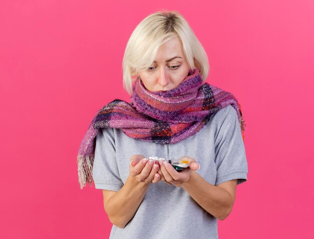 Geschokt jonge blonde zieke Slavische vrouw die sjaal draagt houdt en kijkt naar verpakkingen van medische pillen