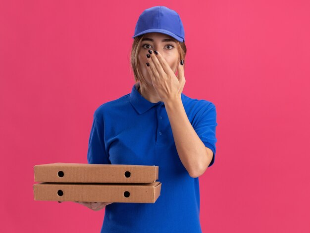 Geschokt jonge mooie levering meisje in uniform legt hand op mond en pizzadozen op roze houdt