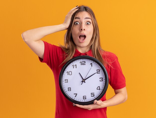 Geschokt jonge mooie vrouw legt hand op het hoofd en houdt klok geïsoleerd op oranje muur