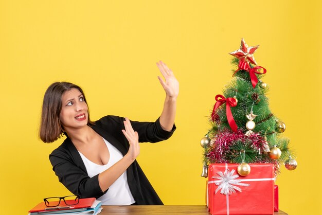 Geschokt jonge vrouw met tien zittend aan een tafel in de buurt van versierde kerstboom op kantoor op geel