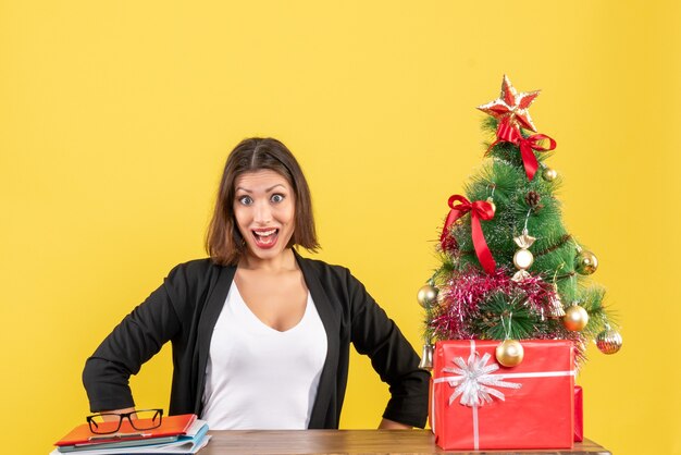 Geschokt jonge vrouw zittend aan een tafel in pak in de buurt van versierde kerstboom op kantoor op geel