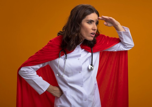 Geschokt kaukasisch superheld meisje in uniform arts met rode cape en stethoscoop houdt palm