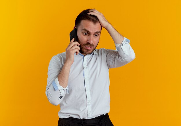 Geschokt knappe man legt hand op hoofd praten over telefoon geïsoleerd op oranje muur