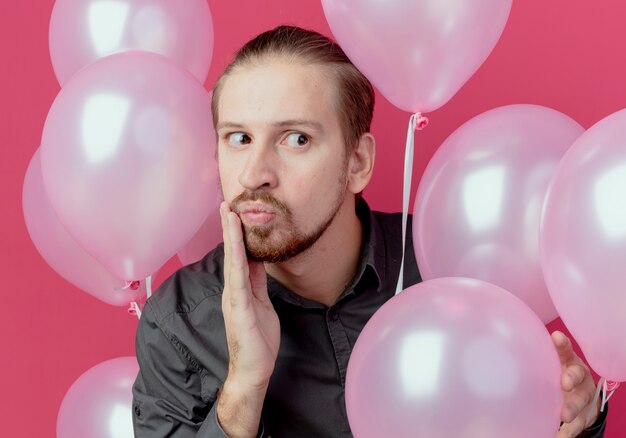 Geschokt knappe man staat met helium ballonnen legt hand op gezicht kijken kant geïsoleerd