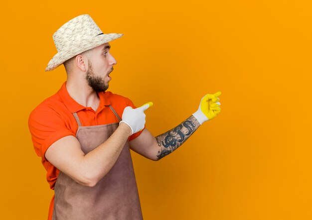 Geschokt mannelijke tuinman tuinieren hoed en handschoenen dragen kijkt en wijst naar de zijkant