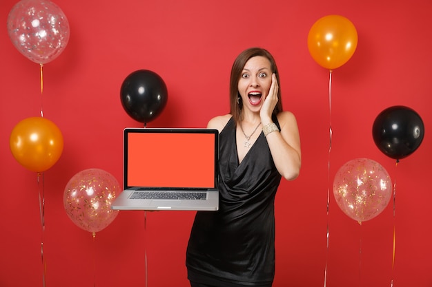 Geschokt meisje in zwarte jurk die hand op gezicht houdt met laptop pc-computer met leeg zwart leeg scherm op heldere rode achtergrondluchtballonnen. gelukkig nieuwjaar, verjaardag mockup vakantie feest concept.