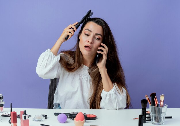 Geschokt mooi meisje zit aan tafel met make-up tools haar kammen praten over telefoon geïsoleerd op paarse muur