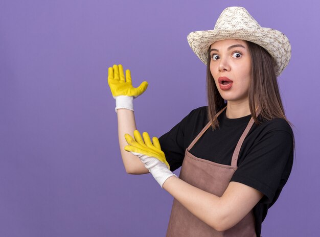 Geschokt mooie blanke vrouwelijke tuinman met tuinhoed en handschoenen die terug wijst met handen geïsoleerd op paarse muur met kopieerruimte