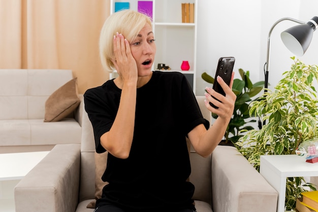 Geschokt mooie blonde Russische vrouw zit op fauteuil hand zetten gezicht kijken naar telefoon in woonkamer