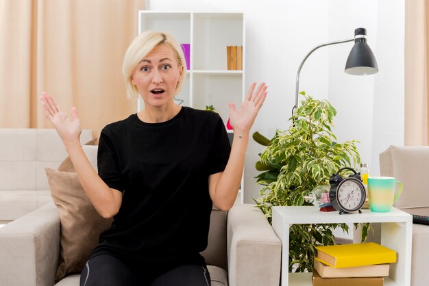 Geschokt mooie blonde Russische vrouw zit op fauteuil handen opheffen in de woonkamer
