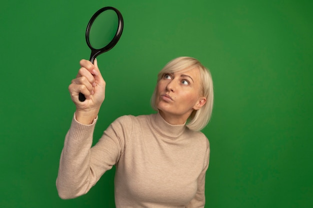 Gratis foto geschokt mooie blonde slavische vrouw houdt en kijkt naar vergrootglas op groen