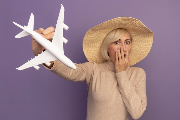 Gratis foto geschokt mooie blonde slavische vrouw met strandhoed legt hand op mond en houdt modelvliegtuig op paars