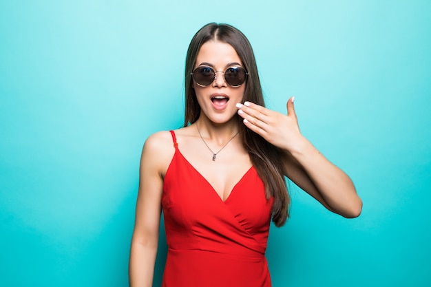 Geschokt mooie jonge vrouw in rode minikleding en zonnebril bedekken mond met hand op blauwe muur.